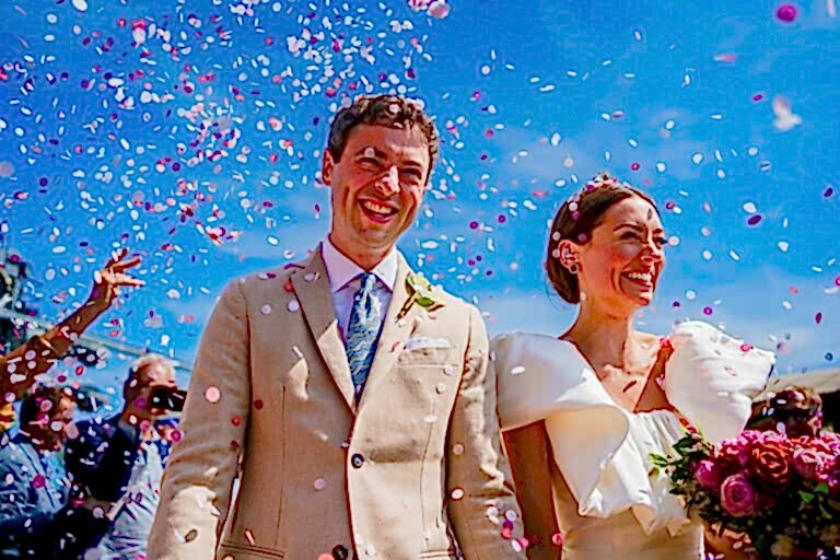 A newlywed couple walks down the aisle outdoors under a bright blue sky. The groom, in a beige suit, and the bride, in a white dress with ruffled sleeves, both smile joyfully as confetti rains down on them. The bride holds a bouquet of red and pink flowers. Image by Documentary Wedding Photographer Lewis Bishop Photography.