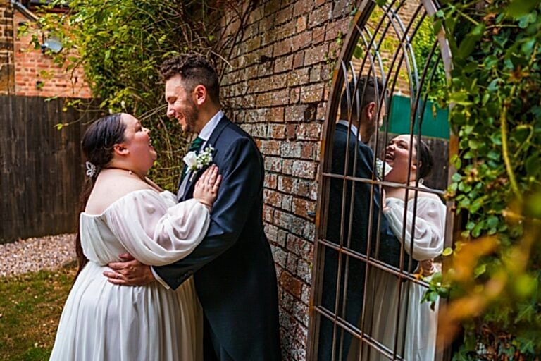 A couple in wedding attire stands close together by a brick wall with greenery, smiling and looking into each other's eyes. Their reflections are visible in the decorative mirror on the wall, adding depth to the romantic scene. Image by Documentary Wedding Photographer Lewis Bishop Photography.