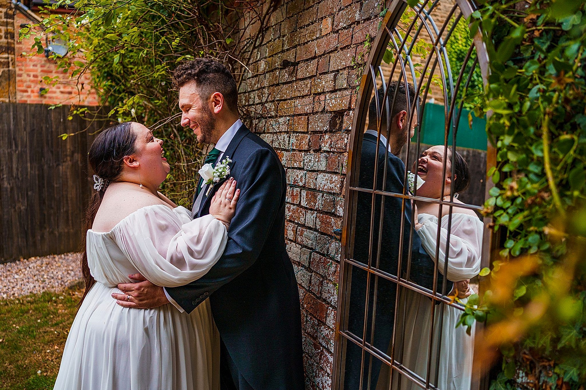 A couple in wedding attire stands close together by a brick wall with greenery, smiling and looking into each other's eyes. Their reflections are visible in the decorative mirror on the wall, adding depth to the romantic scene. Image by Documentary Wedding Photographer Lewis Bishop Photography.