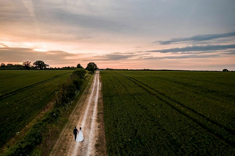 A couple walks hand in hand down a straight dirt road through vast green fields at sunset, with a tree line in the distance under a partly cloudy sky. Image by Documentary Wedding Photographer Lewis Bishop Photography.