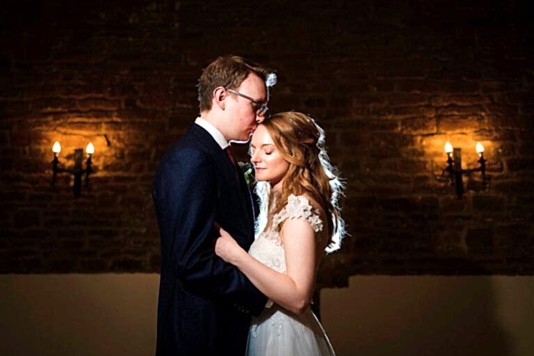 A couple in wedding attire stands close together in a dimly lit room with exposed brick walls and two wall-mounted light fixtures. The groom kisses the bride's forehead while she smiles softly, both illuminated warmly by the ambient lighting. Image by Documentary Wedding Photographer Lewis Bishop Photography.