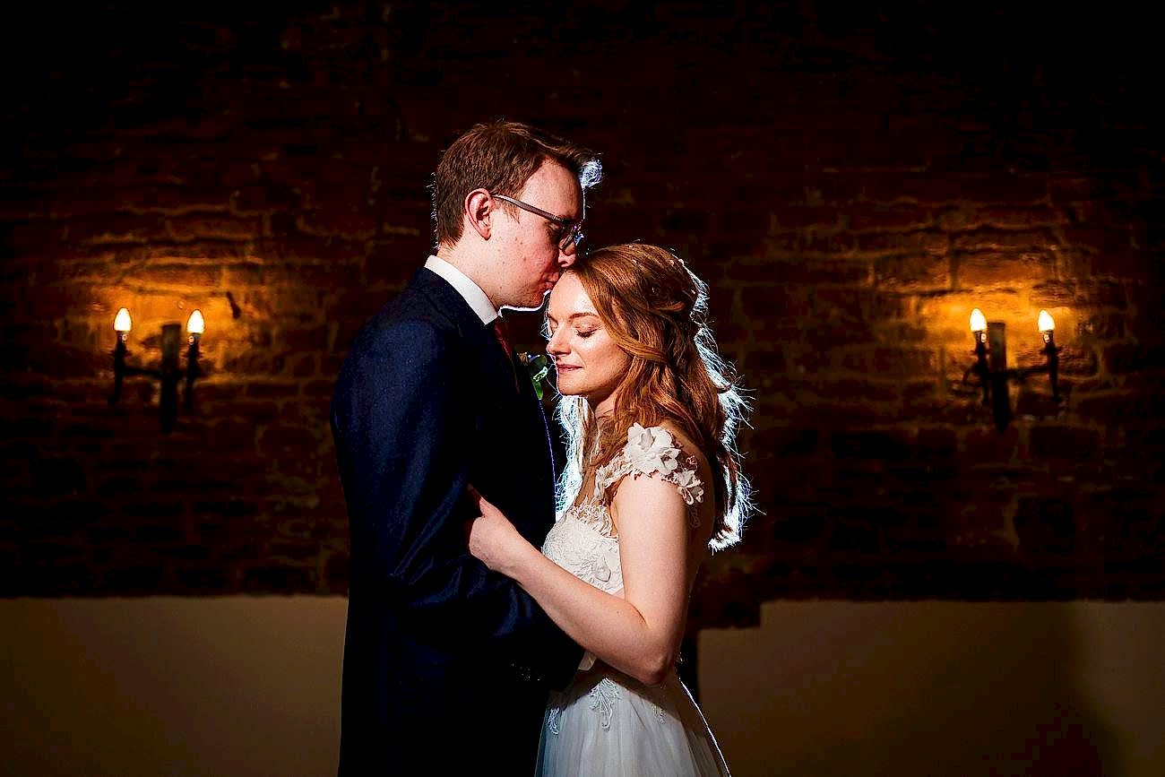 A couple in wedding attire stands close together in a dimly lit room with exposed brick walls and two wall-mounted light fixtures. The groom kisses the bride's forehead while she smiles softly, both illuminated warmly by the ambient lighting. Image by Documentary Wedding Photographer Lewis Bishop Photography.