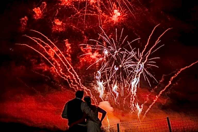 A couple stands close together in front of a fence, gazing at a brilliant fireworks display in the night sky. The fireworks burst in red and white hues, illuminating the smoky sky and creating a festive, romantic atmosphere. Image by Documentary Wedding Photographer Lewis Bishop Photography.