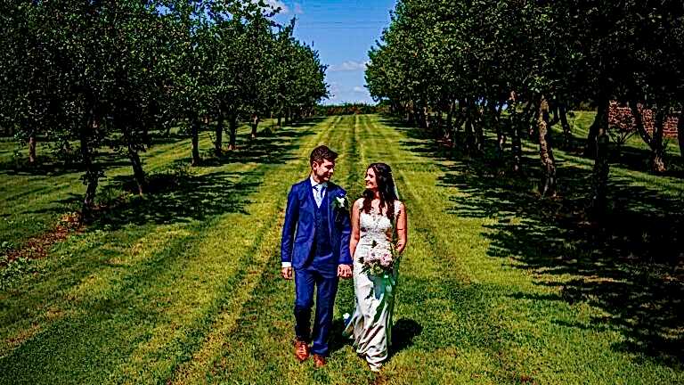 A bride and groom walk hand in hand through a lush, green orchard on a sunny day. The groom is in a blue suit with a white boutonniere, and the bride wears a white wedding dress, holding a bouquet of flowers. Trees line both sides of the path they walk on. Image by Documentary Wedding Photographer Lewis Bishop Photography.