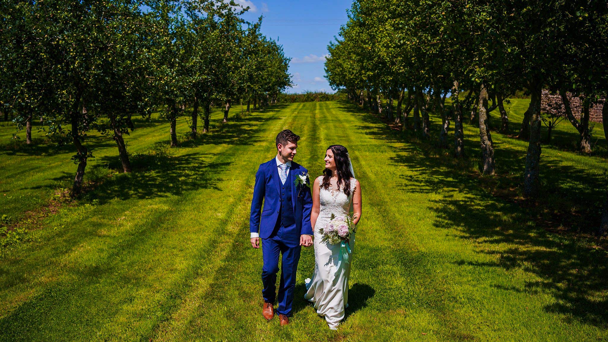 A bride and groom walk hand in hand through a lush, green orchard on a sunny day. The groom is in a blue suit with a white boutonniere, and the bride wears a white wedding dress, holding a bouquet of flowers. Trees line both sides of the path they walk on. Image by Documentary Wedding Photographer Lewis Bishop Photography.