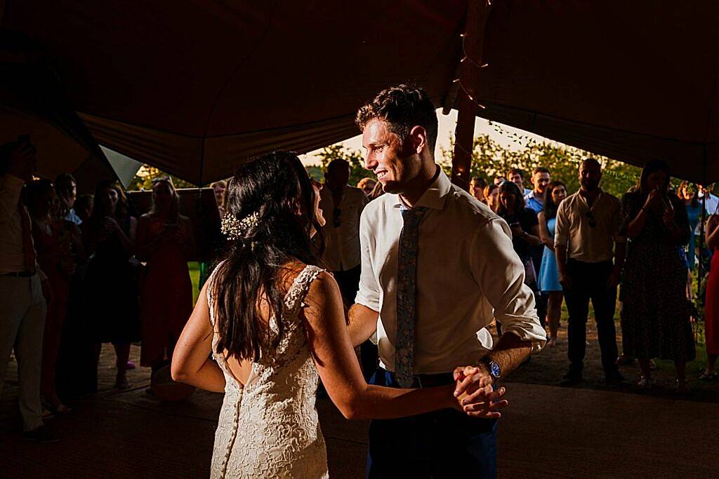 A newlywed couple shares a dance under a canopy tent at sunset. The groom, in a white shirt and blue tie, gazes lovingly at the bride, who is wearing a lace wedding dress with floral hair accessories. Guests stand around them, watching and taking pictures. Image by Documentary Wedding Photographer Lewis Bishop Photography.