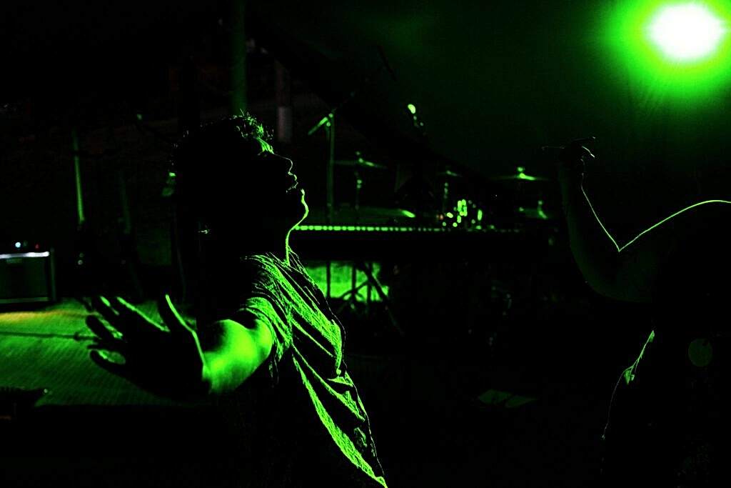 A person stands with arms outstretched, bathed in green stage lighting. The background features musical instruments, including a keyboard and drum set, suggesting a live music performance or a concert setting. Another person is partially visible with a raised hand. Image by Documentary Wedding Photographer Lewis Bishop Photography.