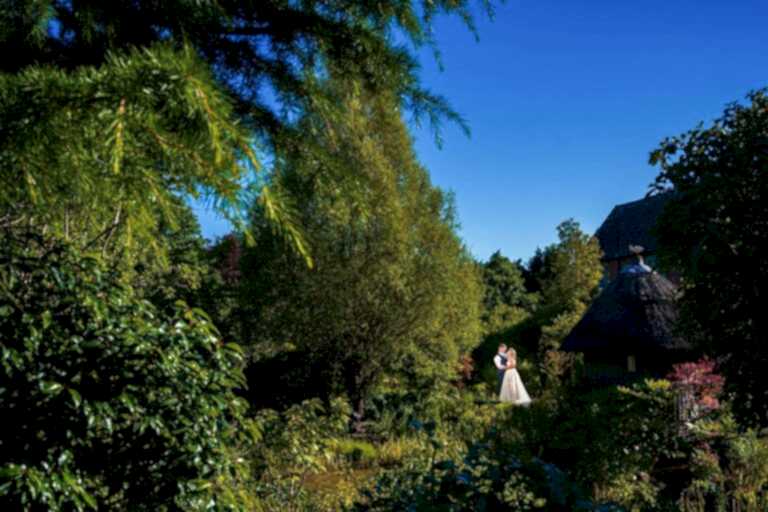 A couple in wedding attire stands close together in an open area surrounded by lush greenery and trees under a clear blue sky. Nearby is a thatched-roof structure partially hidden by the foliage. The scene has a serene, romantic atmosphere. Image by Documentary Wedding Photographer Lewis Bishop Photography.