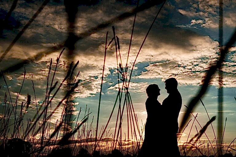 Silhouetted couple standing close together in a field at sunset, surrounded by tall grass, with a dramatic sky of clouds and the setting sun casting a warm glow. Image by Documentary Wedding Photographer Lewis Bishop Photography.