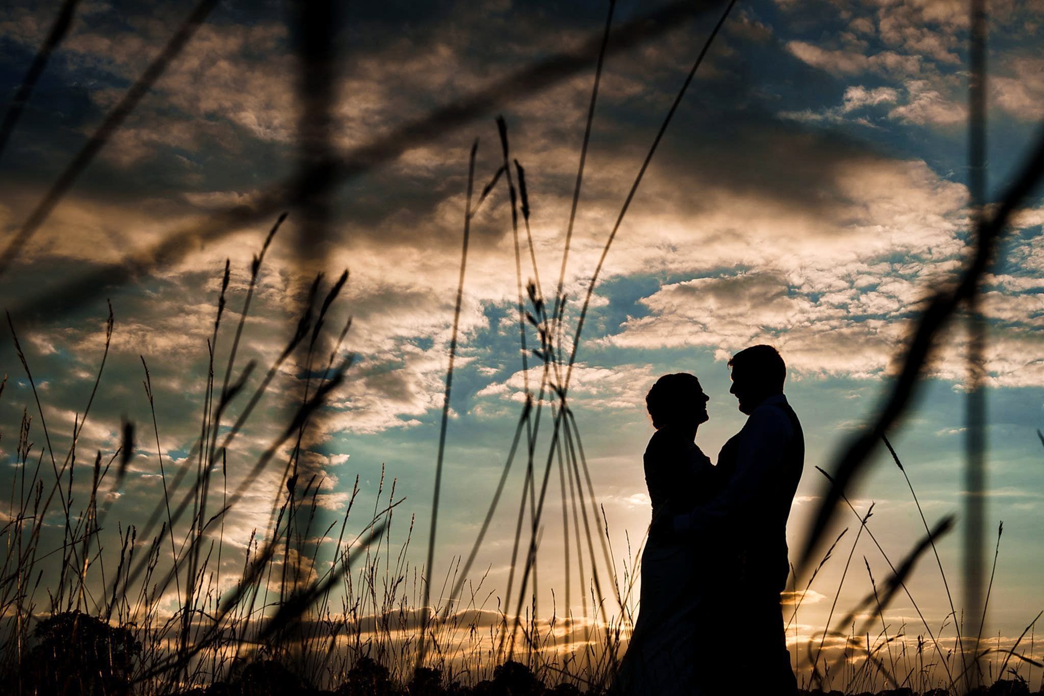 Silhouetted couple standing close together in a field at sunset, surrounded by tall grass, with a dramatic sky of clouds and the setting sun casting a warm glow. Image by Documentary Wedding Photographer Lewis Bishop Photography.