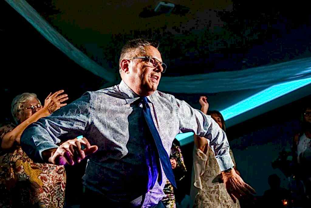 A man wearing a patterned shirt and tie dances energetically at a party. He appears joyful and is surrounded by other people who are also enjoying the festive atmosphere. Blue lighting and draped fabric add to the celebratory mood of the scene. Image by Documentary Wedding Photographer Lewis Bishop Photography.