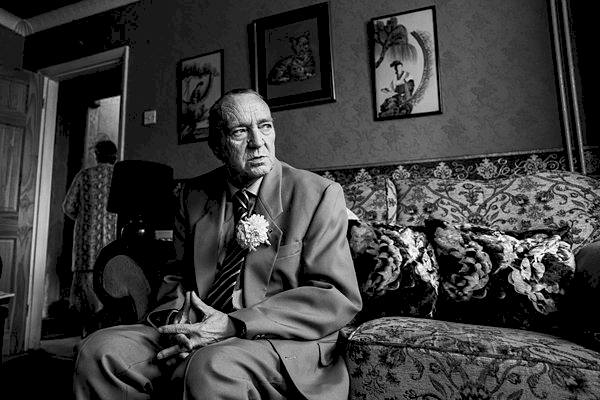 Elderly man in suit sitting on patterned sofa, contemplative.