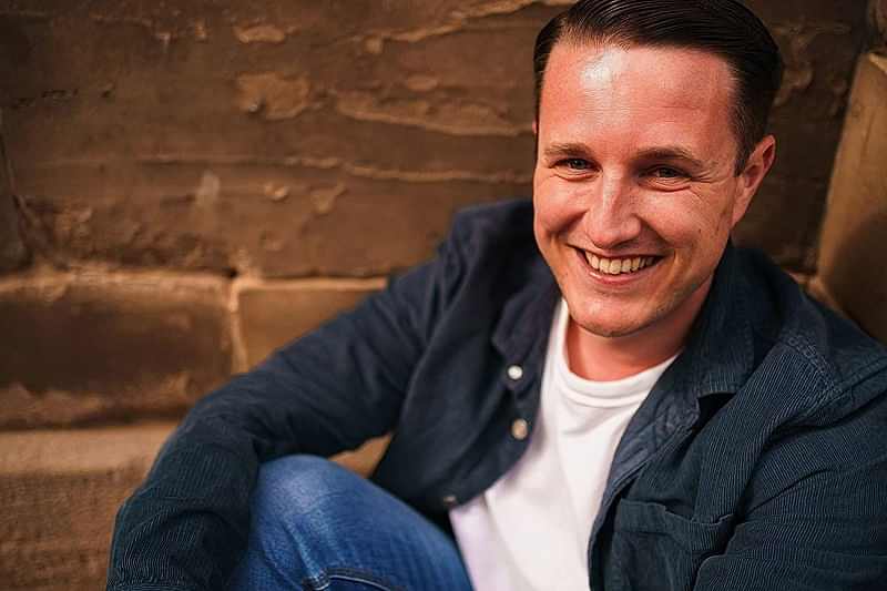 Smiling man sitting casually against brick wall.