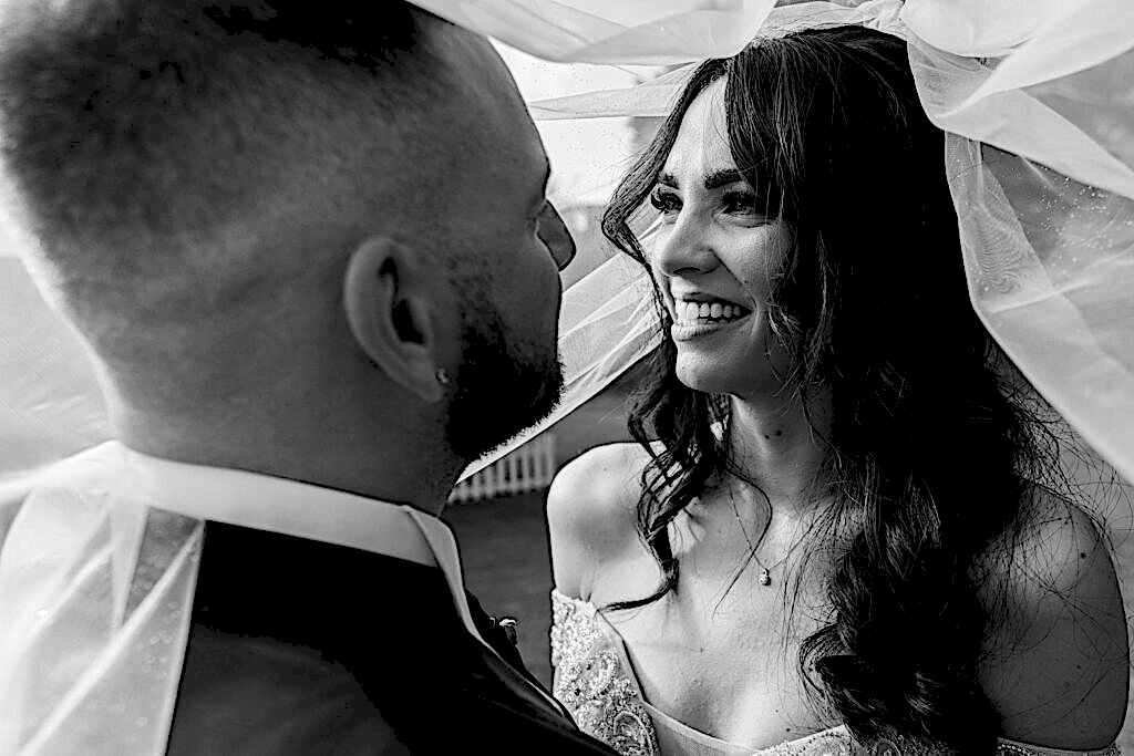 Bride and groom close-up under veil, smiling, black and white.