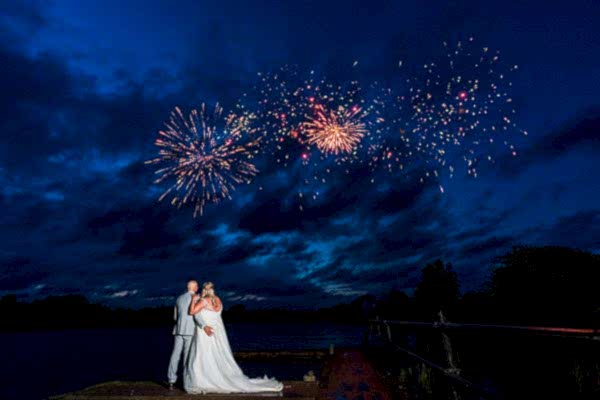 Couple kissing under fireworks at night by lake.