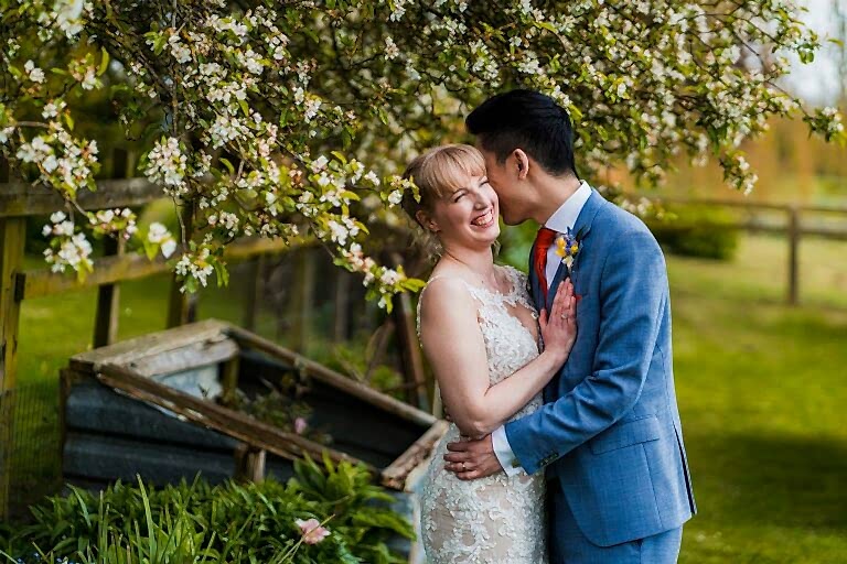 A joyful couple poses under a blooming tree. The bride, in an ornate white dress, smiles brightly as the groom, in a light blue suit, kisses her cheek. They stand in a lush garden with wooden structures and greenery in the background. Image by Documentary Wedding Photographer Lewis Bishop Photography.