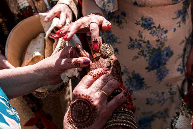 A close-up image showing multiple hands adorned with intricate henna designs and jewelry. One person is adjusting a golden watch on another's wrist, while others delicately gesture around. The scene is vibrant, with floral clothing patterns in the background. Image by Documentary Wedding Photographer Lewis Bishop Photography.