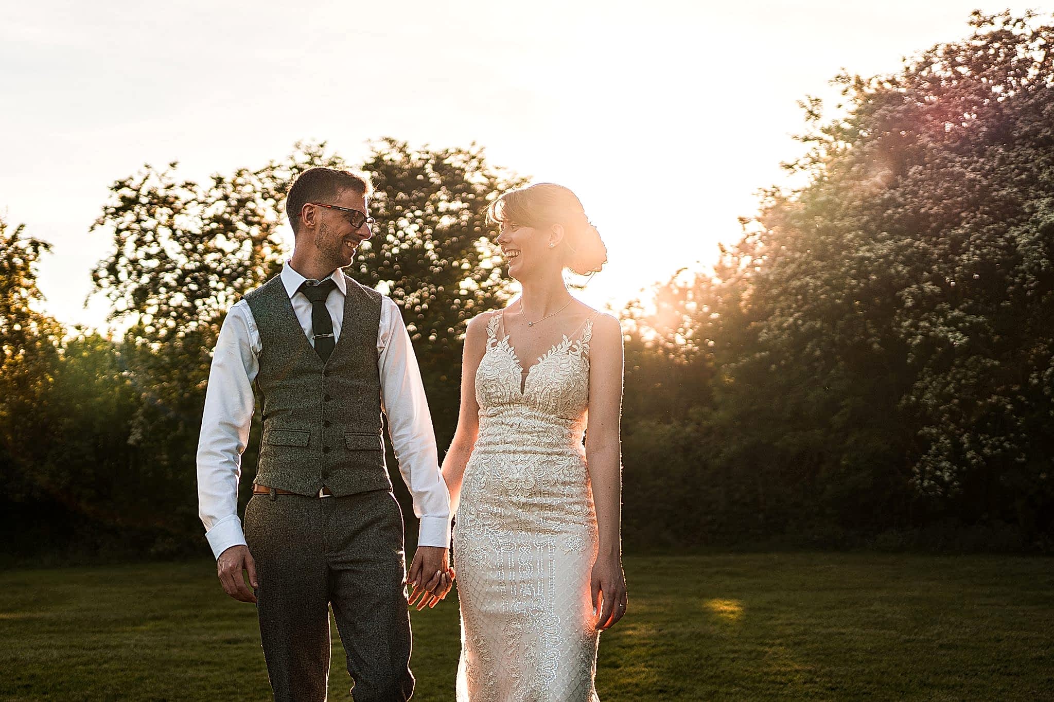 A couple, dressed in wedding attire, walk hand in hand outdoors during sunset. The bride wears a fitted white lace gown, and the groom is in a vest and tie. They smile at each other, with trees and a golden sky in the background, creating a romantic ambiance. Image by Documentary Wedding Photographer Lewis Bishop Photography.
