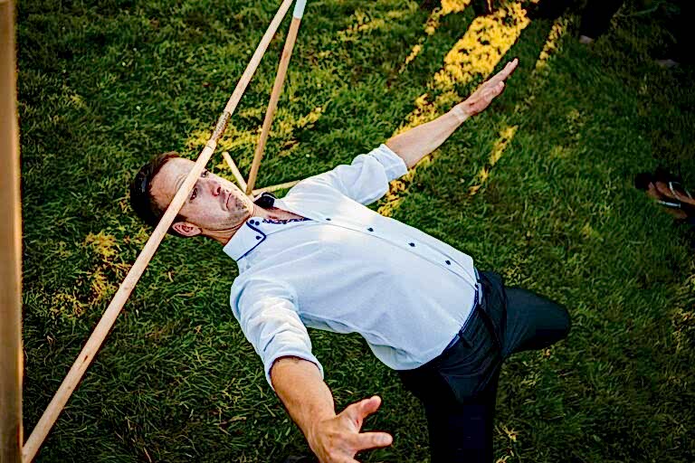 A man in a white shirt and black pants leans backward under a limbo stick on a grassy area, balancing carefully with his arms outstretched. Sunlight casts shadows, and a pair of shoes can be seen on the grass in the background. Image by Documentary Wedding Photographer Lewis Bishop Photography.