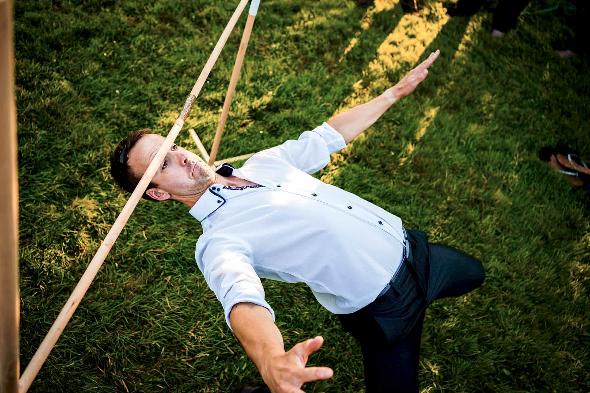 A man in a white shirt and black pants leans backward under a limbo stick on a grassy area, balancing carefully with his arms outstretched. Sunlight casts shadows, and a pair of shoes can be seen on the grass in the background. Image by Documentary Wedding Photographer Lewis Bishop Photography.