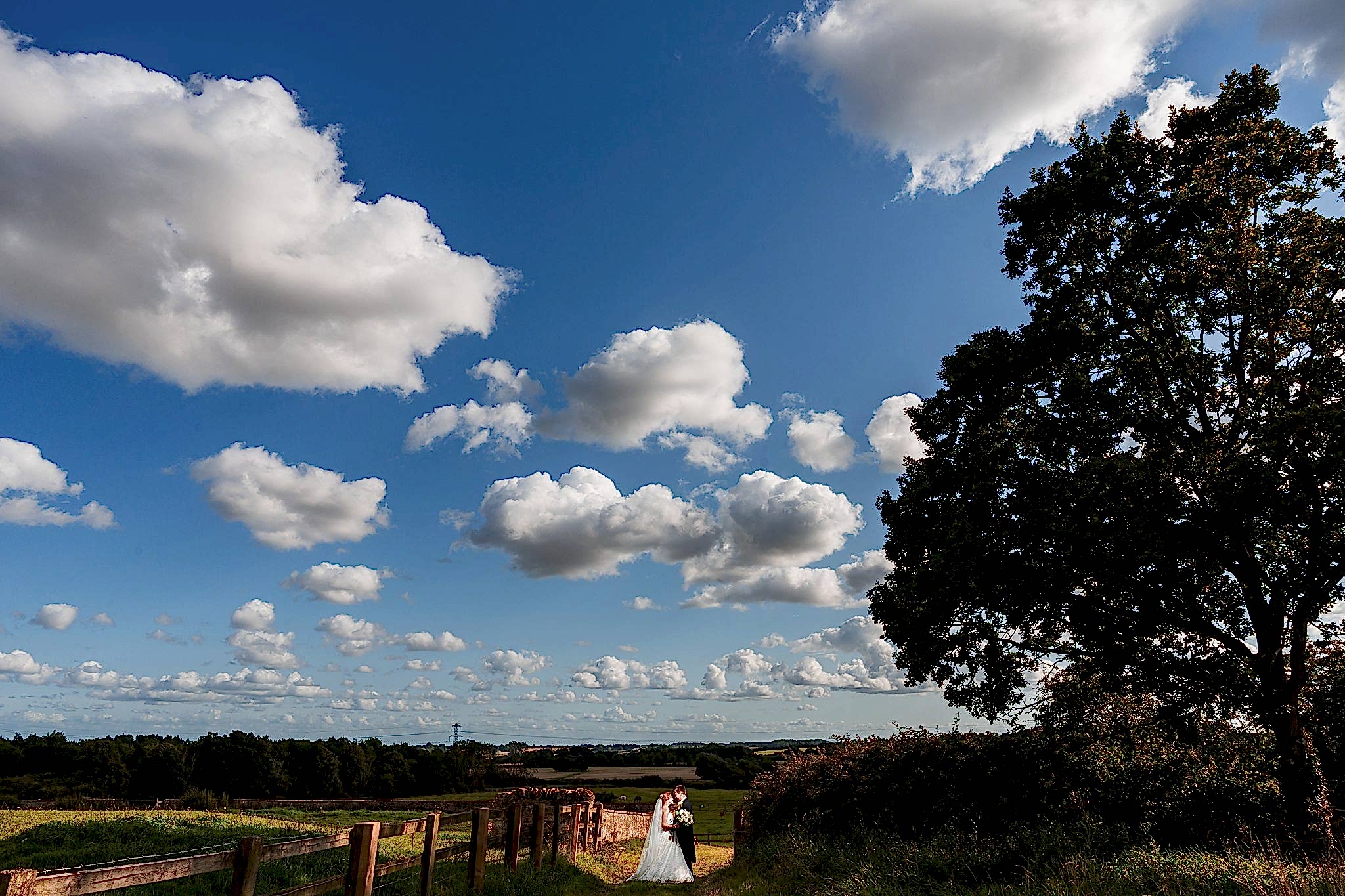 A couple dressed in wedding attire poses under a vast, clear blue sky dotted with fluffy white clouds. They stand beside a wooden fence in a scenic countryside landscape with lush green fields, a tall tree on the right, and distant trees on the horizon. Image by Documentary Wedding Photographer Lewis Bishop Photography.