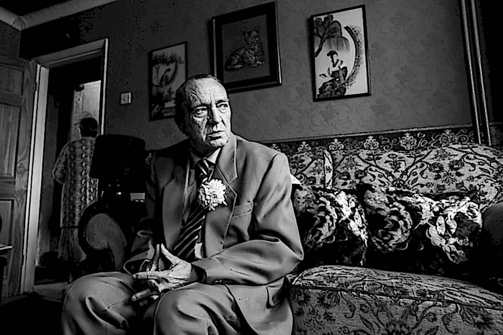 Elderly man in suit, seated on floral sofa.
