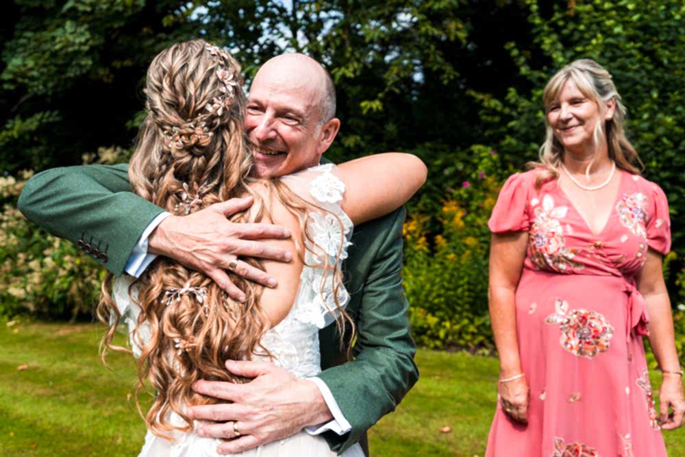 Bride hugging man with woman in pink dress smiling.