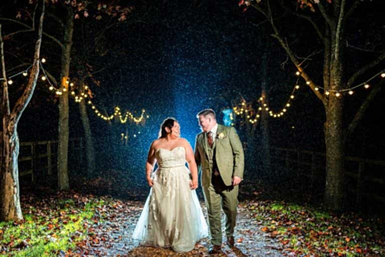Bride and groom walking under string lights in rain.