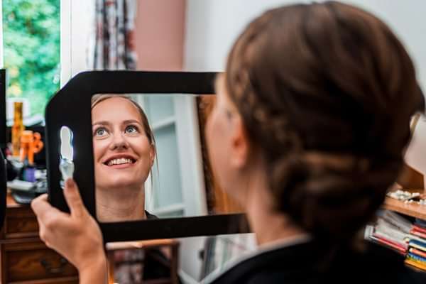 Woman smiling at reflection in handheld mirror.