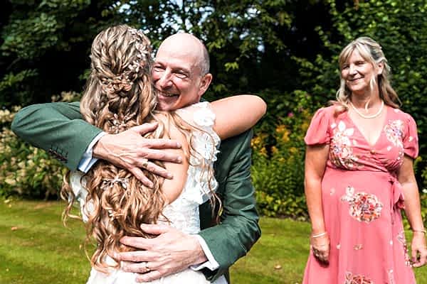 Bride hugging older man outdoors, smiling woman nearby.