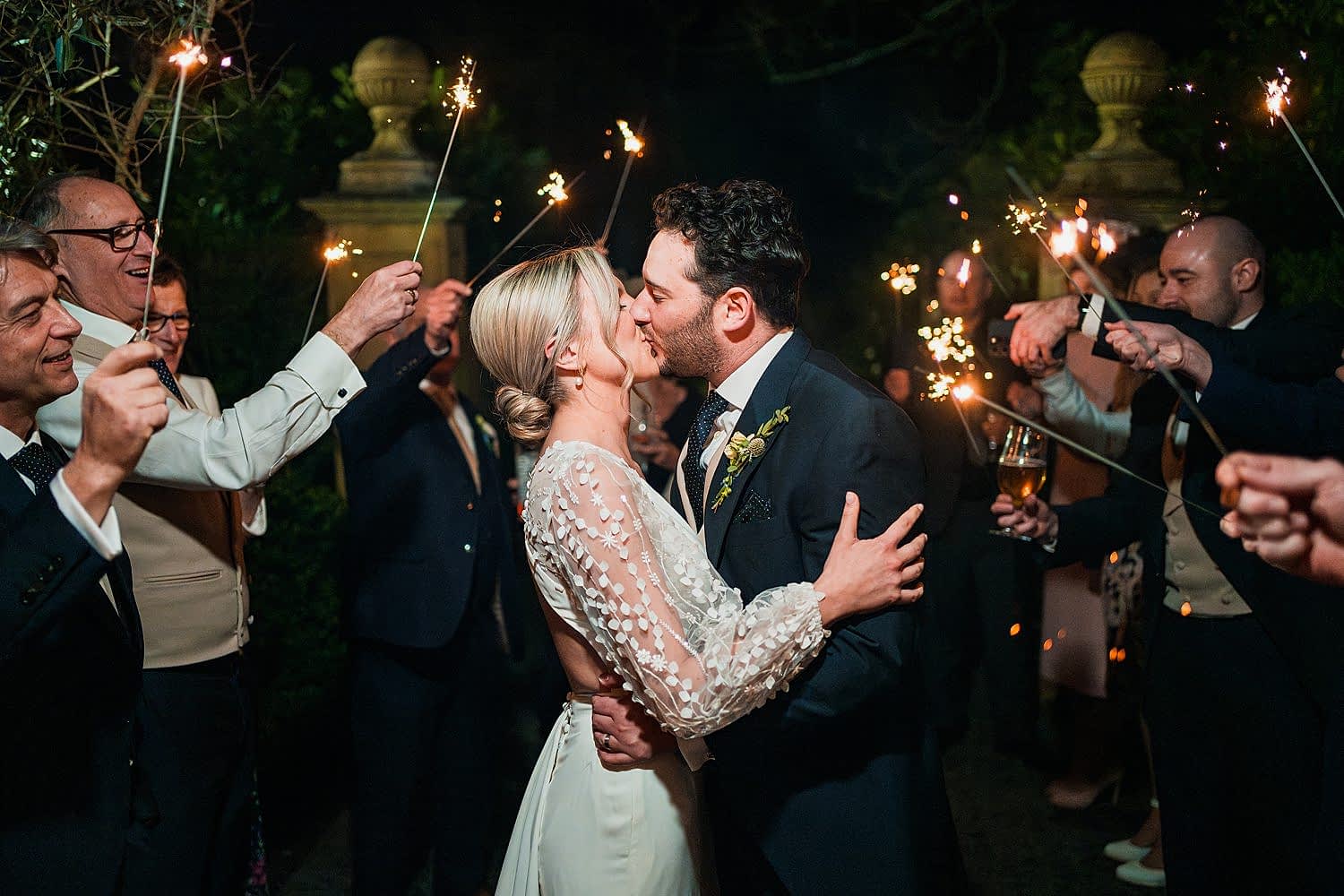 Bride and groom kiss surrounded by sparklers.