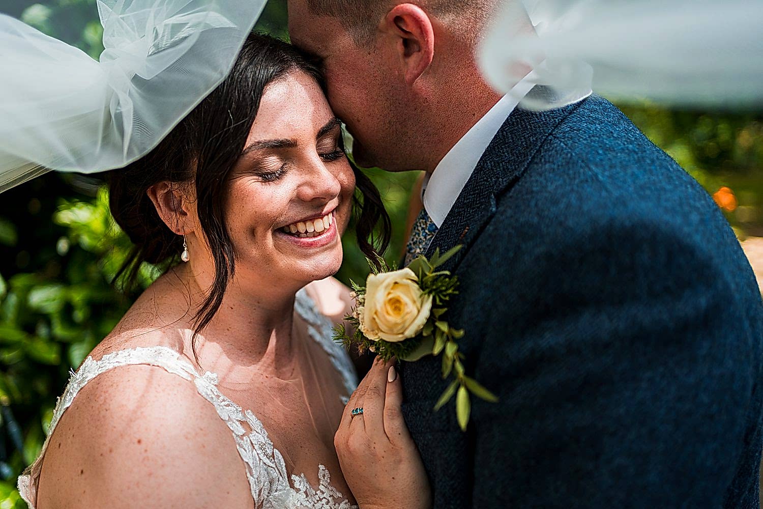 Smiling bride and groom under veil with bouquet.